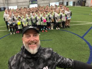 Large group of youth soccer players in white-green uniforms posing for a team photo inside an indoor soccer facility, with the coach taking a selfie in the foreground.